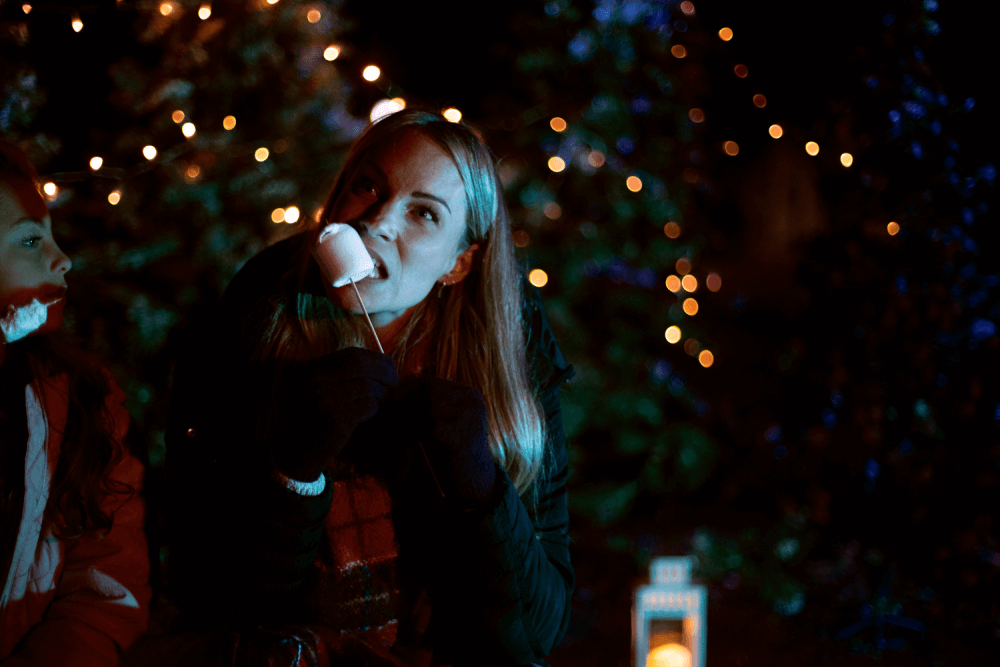A woman eats a toasted marshmallow on a stick around a fire pit at christmas