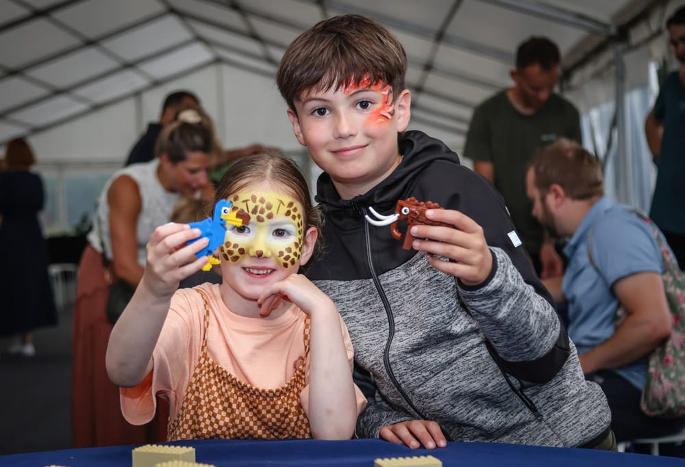 Two children with their faces painted hold up the Dodo and Mammoth they made from toy bricks