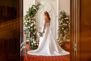 A bride holding a bouquet of flowers stands in front of a flower arch, during her wedding at Marwell Hall
