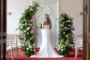 A bride stands in front of a floral arch in the Long Room in Marwell Hall