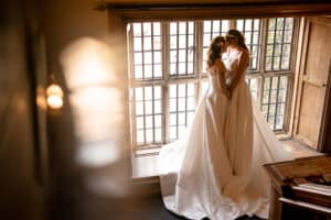 Two brides embrace in front of a large window in Marwell Hall